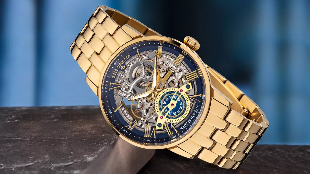 Gold skeleton watch with blue accents, visible gears, and matching gold metal bracelet, displayed on a dark stone surface with a blurred blue background.