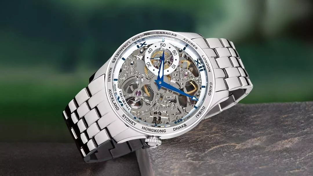 Tufina stainless steel skeleton watch with blue hands, world time bezel, and metal link bracelet, displayed on a stone surface with blurred green background.