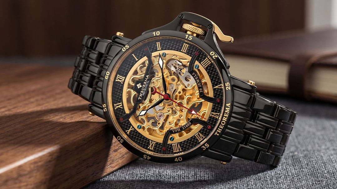 A luxury skeleton watch with a black link bracelet, black bezel, and a gold-tone mechanical dial showing the inner gears. The watch is resting on a dark wooden block over a gray textured surface, with a blurred book in the background.